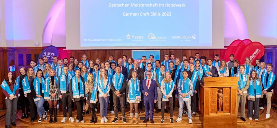 Auf dem Bild sind 81 junge Menschen mit blauen Schals zu sehen, auf denen das Logo von "Das Handwerk" ist. dazwischen stehen Senator Ties Rabe und Handwerkskammerpräsident Hjalmar Stemmann.