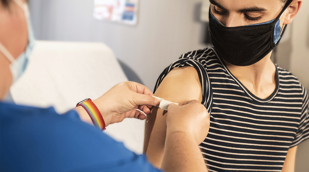 Das Bild zeigt eine Person die im rechten Oberarm von einer anderen Person ein Pflaster aufgeklebt bekommt. Vermutlich passiert dies nach einer Injektion für eine Impfung.
