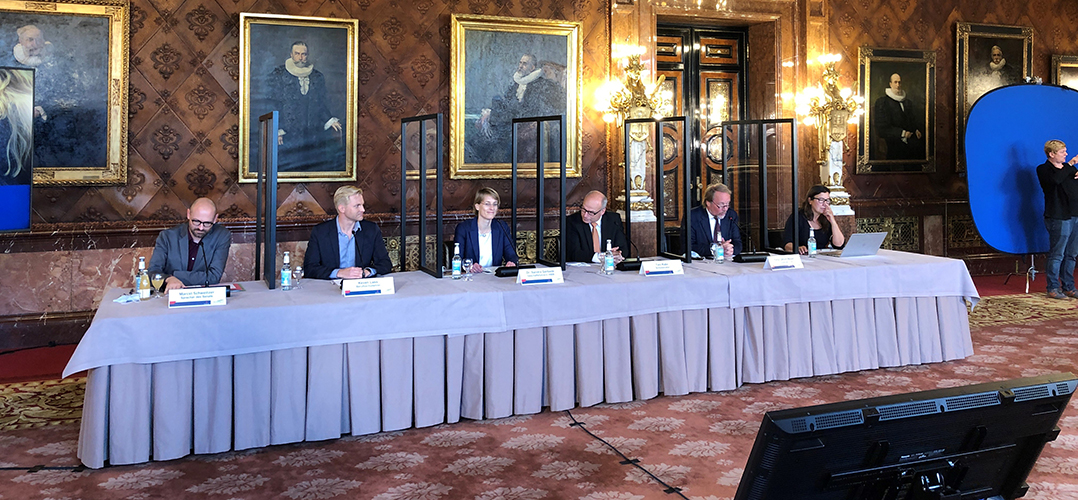 Das Bild zeigt sechs Personen an einem langen Tisch in einem festlichen Raum, vermutlich dem Hamburger Rathaus. Die Personen haben Namensschilder und Mikrofone vor sich, so dass es nach einer Pressekonferenz aussieht.