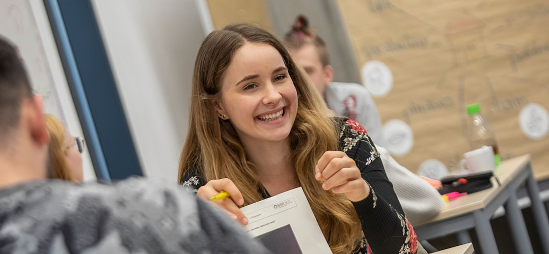 Das Bild zeigt eine lächelnde Person, die sich scheinbar in einem Klassenraum befindet und einen Stift und ein Blatt Papier in der Hand hält.