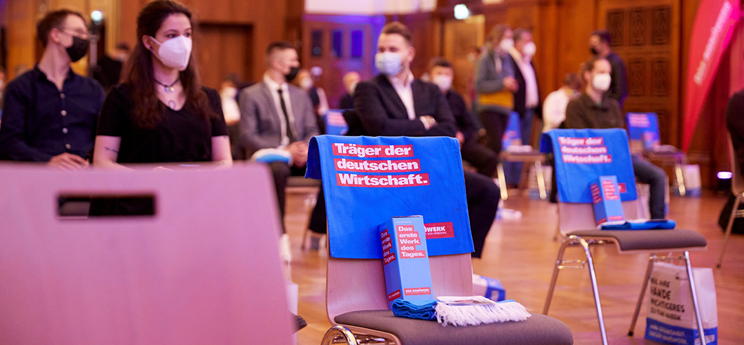 Das Bild zeigt zwei Stühle im Vordergrund, auf denen eine blaue Tasche und eine blaue Box sowie ein blauer Schal liegen. Auf der Tasche steht "Träger der deutschen Wirtschaft". Auf der blauen Box steht:" DAs erste Werk des Tages. Das Handwerk". Im Hintergrund sind mehrere Personen in Anzügen und mit Atemschutzmasken zu sehen.