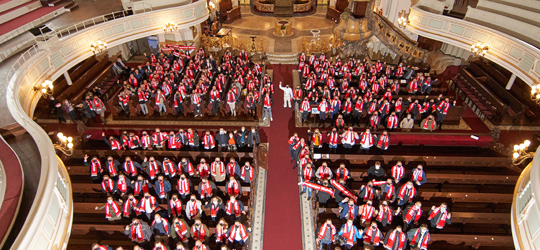 Das Bild zeigt eine große Gruppe von Personen von oben aufgenommen. Der Raum scheint ein festlicher Raum zu sein, mit rotem Teppich ausgekleidet. Die Personen tragen rote Schals mit weißer Aufschrift.