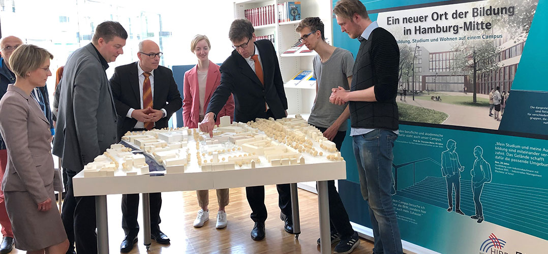 Auf dem Bild sind sieben Personen zu sehen, die vor einem Modell stehen. Das Modell zeigt mehrere Modellgebäude und ist aus Holz. Im Hintergrund ist ein Plakat auf dem steht: "Ein neuer Ort der Bildung in Hamburg-Mitte". Der Raum ist von Tageslicht erhellt.