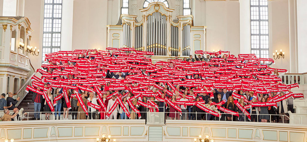 Das Bild zeigt eine größere Gruppe von Personen mit roten Schals, die vor einer großen Orgel stehen. Der Raum ist lichtdurchflutet und ist in einer großen Hamburger Kirche, dem Michel.