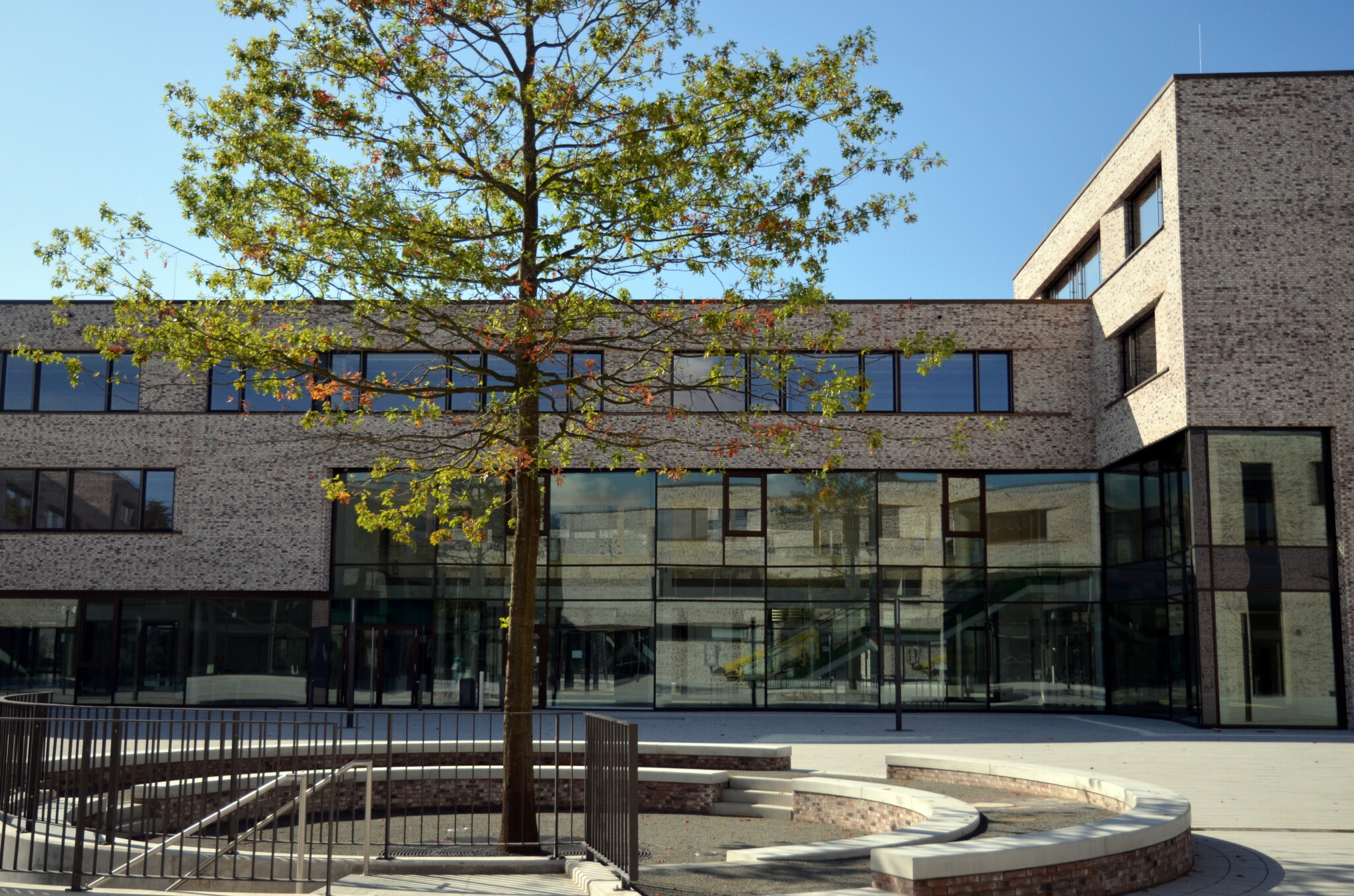 Das Foto zeigt im Hintergrund die BS01, ein 3-geschossiges modernes Gebäude aus braunem Backstein mit großer Fensterfront und Flachdach. Im Vordergrund ist eine runde, aus Steinen gemauerte Bank, die um einen Baum herum reicht.