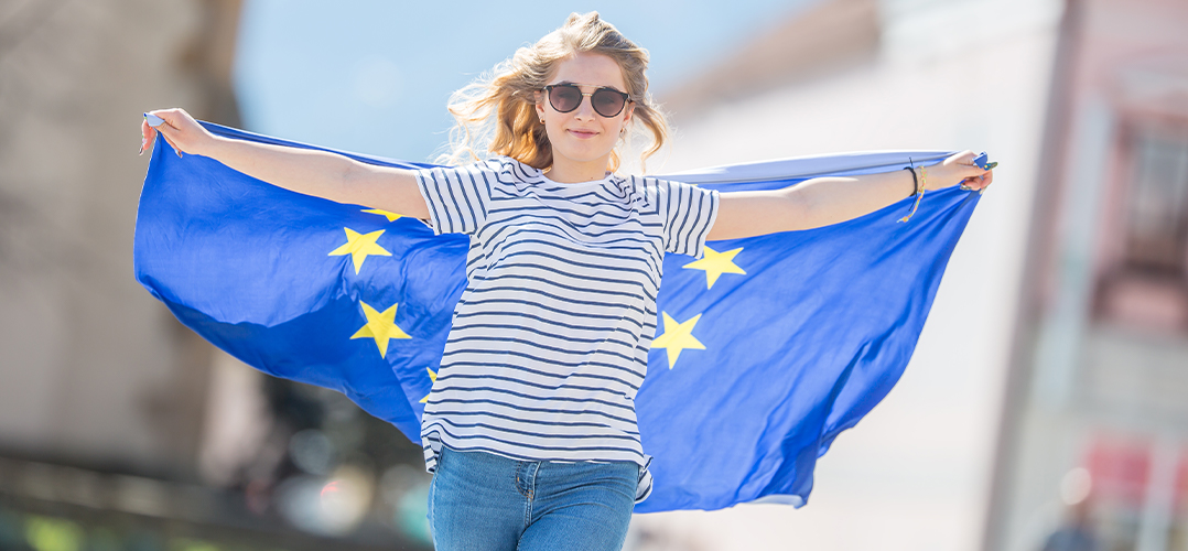 Das Bild zeigt eine weibliche Person, die eine blaue Flagge mit gelben Sternen hält, die Flagge der Europäischen Union (EU). Die Person trägt ein gestreiftes Oberteil und Jeans. Der Hintergrund ist unscharf, aber es sieht nach einem städtischen Umfeld aus mit Gebäuden und klarem Himmel.