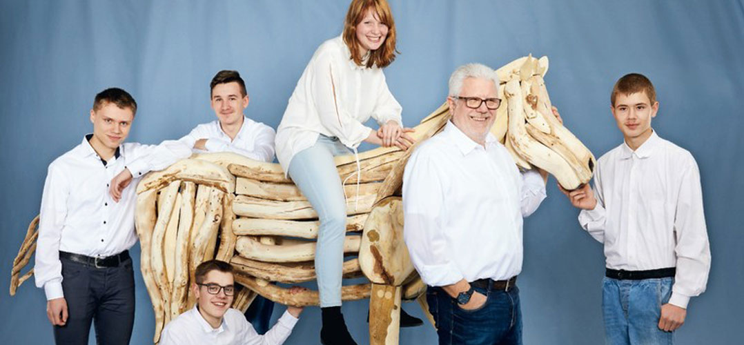 Das Bild zeigt fünf Personen und ein Pferd vor einem blauen Hintergrund. Sie tragen alle weiße Oberteile und dunkle Hosen oder Jeans, außer einer Person, die eine hellblaue Hose trägt. Das Pferd ist ein lebensgroßes Modell aus Holz.