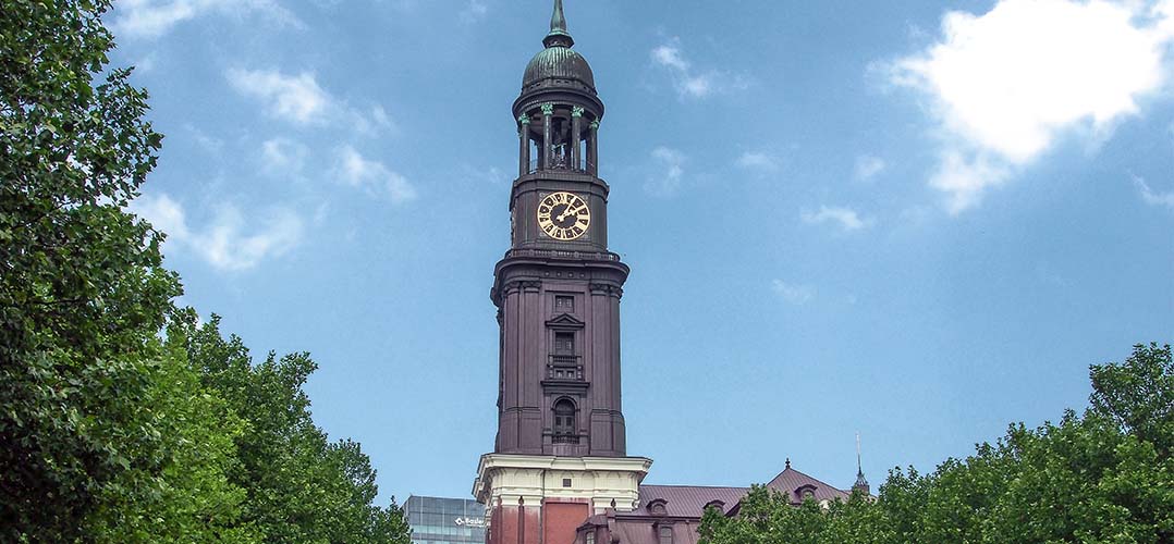 Das Bild zeigt einen hohen Turm mit einer Uhr, der von Bäumen und einem blauen Himmel mit Wolken umgeben ist. Der Turm hat ein historisches Design und ist Teil eines größeren Gebäudes, die Sankt Michaelis Kirche in Hamburg, auch der "Michel" genannt. Der Hauptfokus des Bildes liegt auf einem hohen, dunkelbraunen Turm mit einer großen Uhr nahe der Spitze. Der Turm verfügt über architektonische Details, die auf ein historisches oder traditionelles Design hinweisen. Die Uhr an der Vorderseite des Turms ist groß und deutlich sichtbar; sie zeigt die Zeit an. Grüne Bäume umgeben den unteren Teil des Turms, wobei ihre Blätter einen Kontrast zum dunklen Stein des Gebäudes bilden. Der Himmel im Hintergrund ist blau mit weißen Wolken, was dem Bild eine helle und luftige Atmosphäre verleiht.