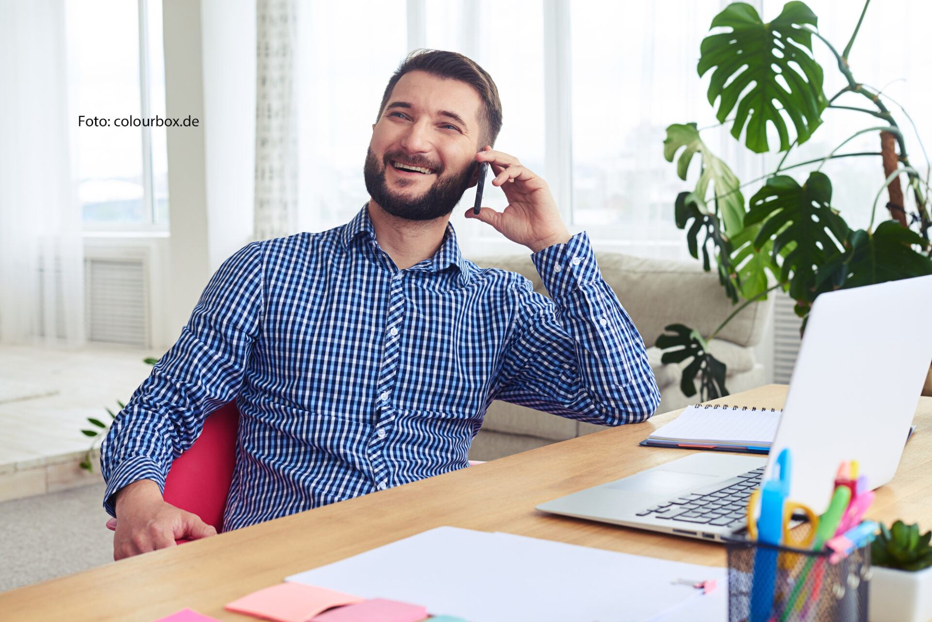 Das Bild zeigt einen bärtigen Mann, vielleicht Mitte 30 Jahre alt, der lachend telefonierend an einem Schreibtisch sitzt. Auf dem Schreibtisch steht ein Notebook.