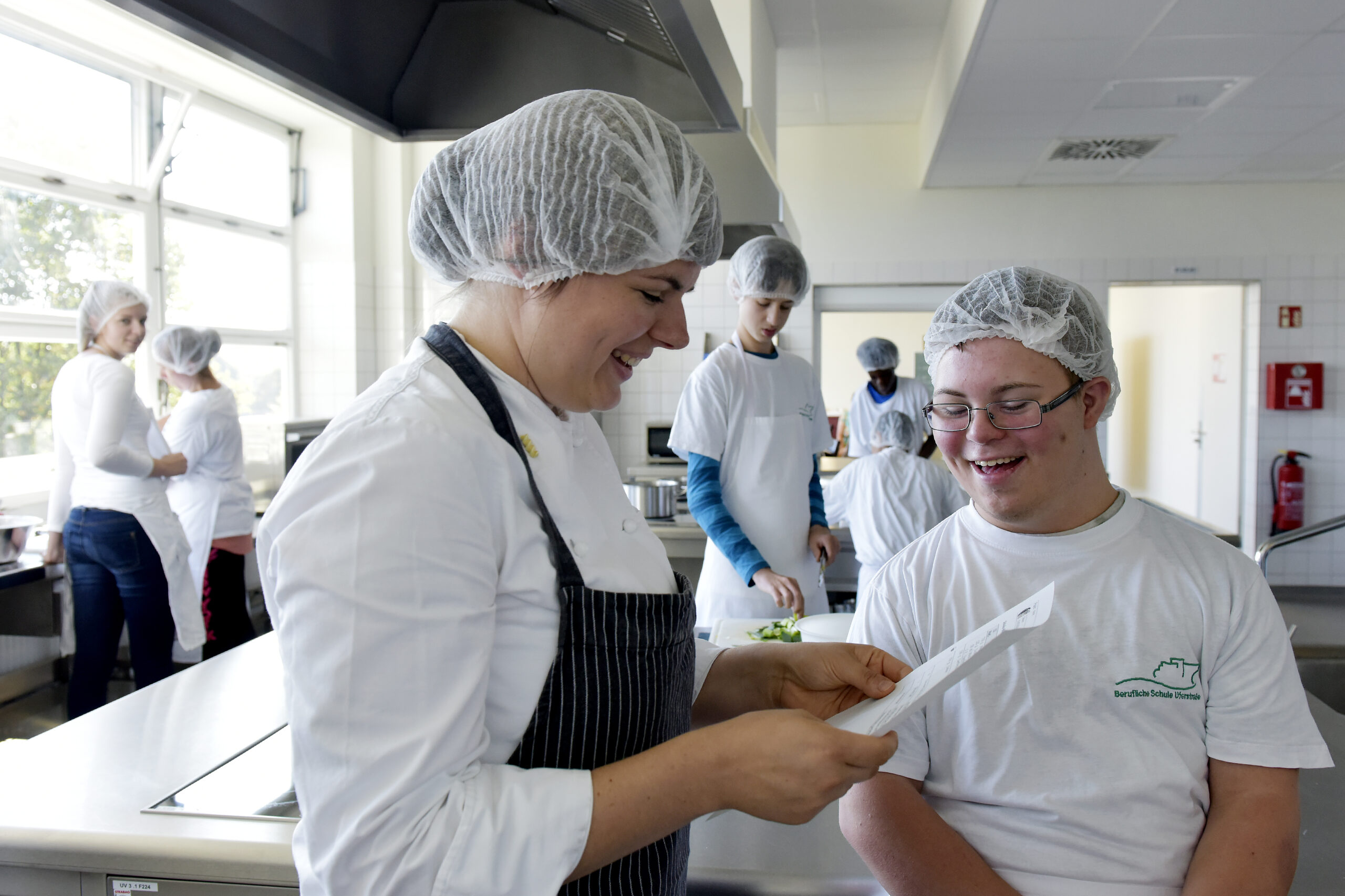 Das Foto zeigt eine Küche mit 5 jungen Menschen mit weißen Kochschürzen im Hintergrund. Im Vordergrund steht links eine junge Frau mit blauer Kochschürze und weißer Haarhaube, die einen Zettel in der Hand hält und einem jungen Mann mit Brille, der lachend auf diesen Zettel schaut.