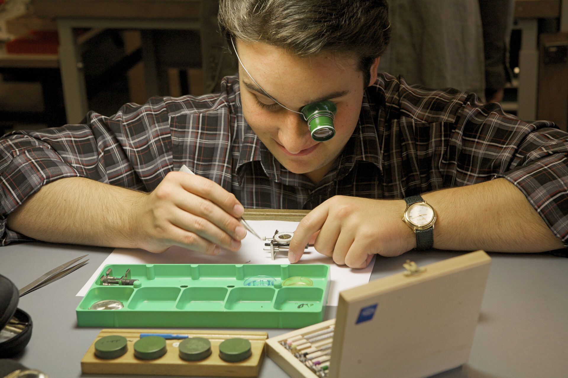 Das Bild zeigt einen sitzenden jungen Mann beim reparieren eines kleinen Uhrwerkes. Er hat eine Lupe vor dem linken Auge befestigt.