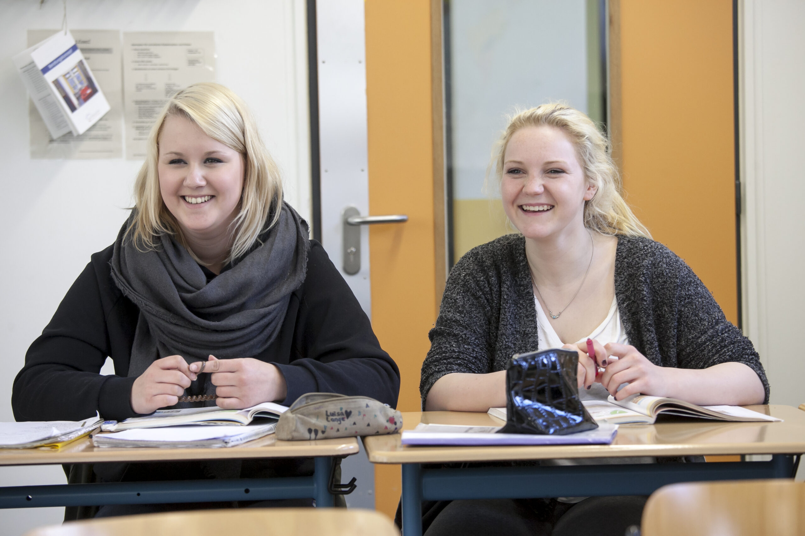 Das Bild zeigt 2 junge blonde Frauen, die beide lächelnd an einem kleinen Tisch sitzen und vor sich ein Schulbuch aufgeschlagen haben.