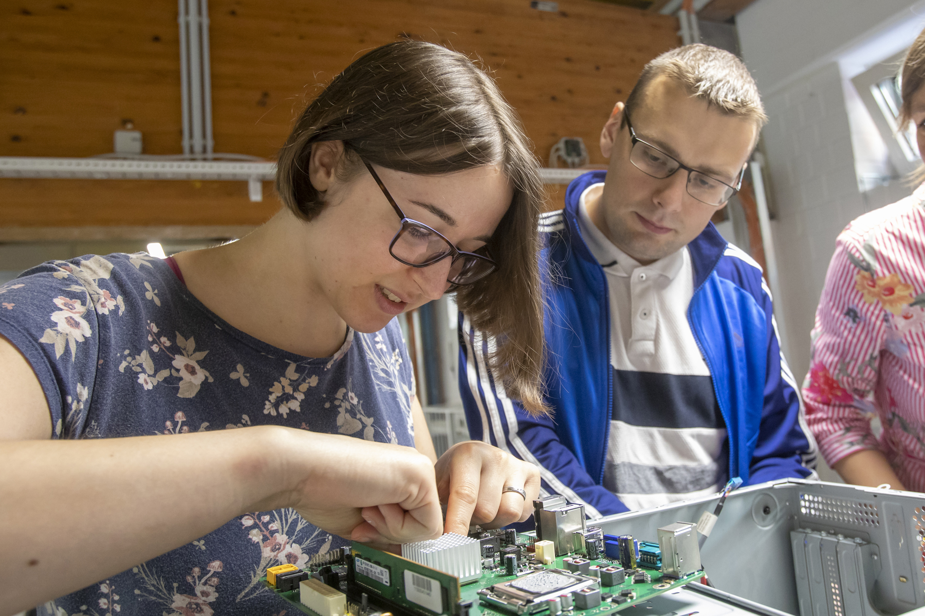 Das Bild zeigt eine junge dunkelhaarige Frau in geblümten blauen T-Shirt bei der Arbeit an einem elektronischen Schaltkreis. Ein junger Mann mit Brille rechts neben ihr schaut dabei zu.