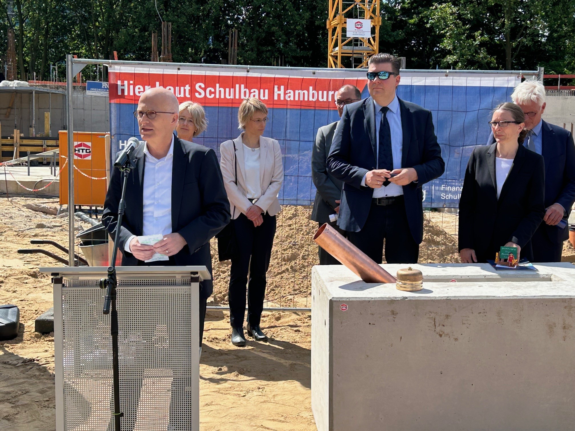 Das Bild zeigt 7 Personen, 4 Männer und 3 Frauen, die vor einer Baustelle stehen. Die linke Person spricht an einem Stehpult in ein Mikrophon.