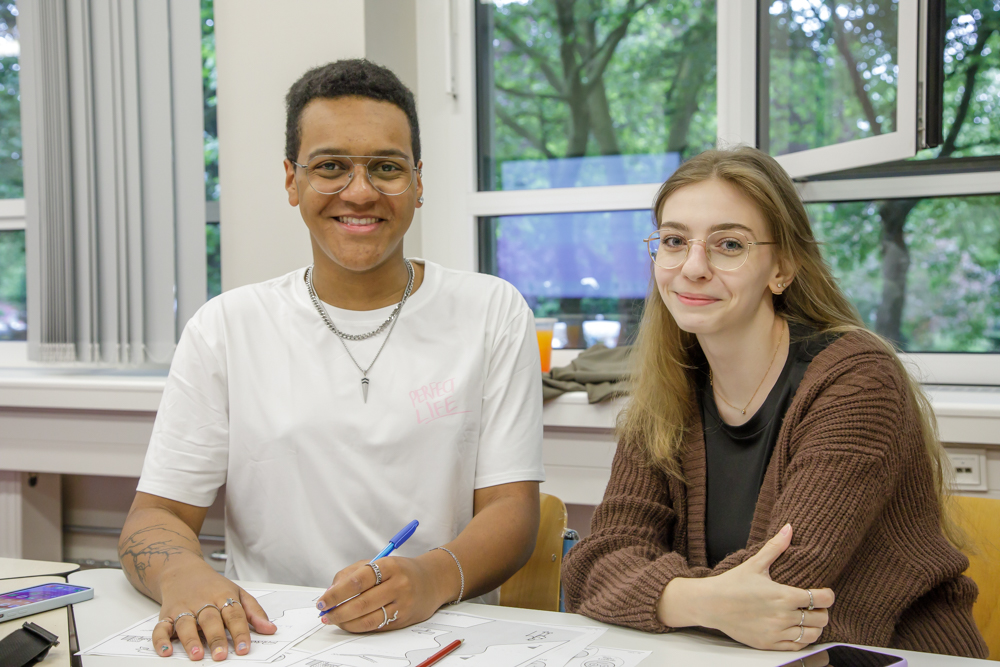 Das Bild zeigt 2 junge Menschen sitzend an einem Tisch. Links ein junger Mann im weißen T-Shirt, rechts eine junge langhaarige Frau in braunem Oberteil