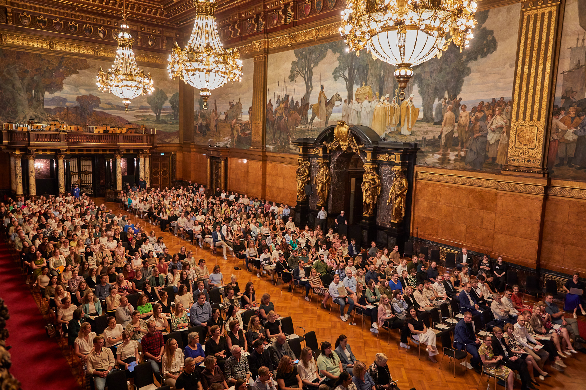 Das Bild zeigt mehrere hundert junge Menschen, die in Reihen auf Stühlen sitzen. Die Umgebung ist ein Festsaal mit hohen Wandgemälden und goldenen Ornamenten und Figuren an den Wandseiten.