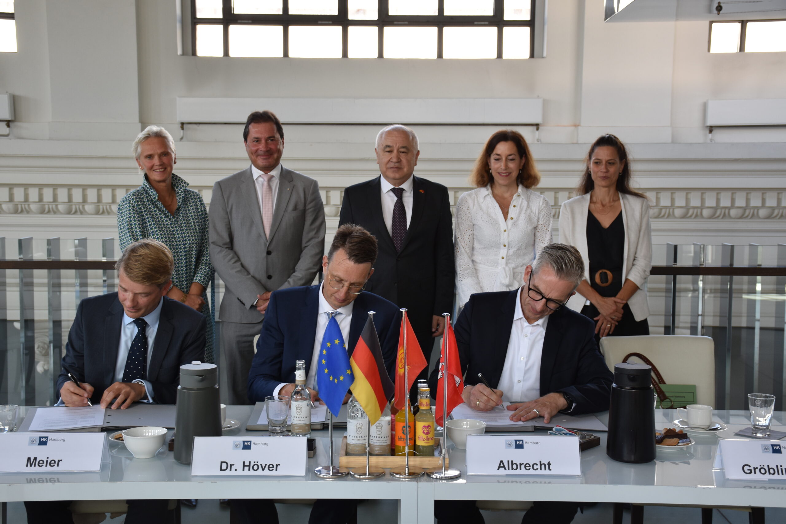 Das Foto zeigt 8 Personen. 3 Männer in blauen Anzügen sitzen an einem Tisch und unterschreiben ein Blatt Papier. Dahinter stehen 5 Personen, 3 Frauen und 2 Männer, die sehr formell nebeneinander stehen und lächelnd schauen