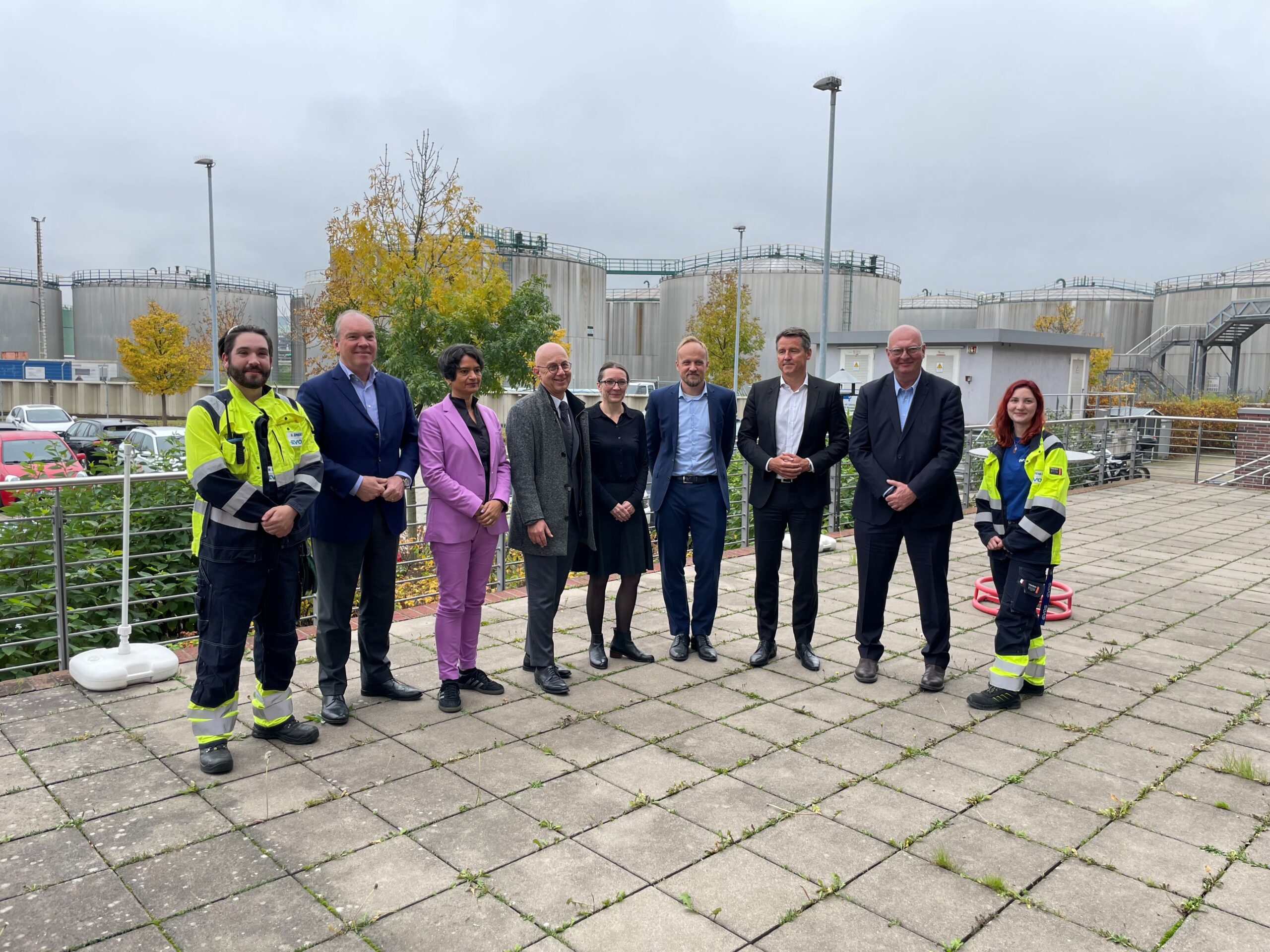 Das Foto zeigt 9 Personen, die auf einer gefliesten Terasse stehen, 6 Männer und 3 Frauen. Alle stehen in einer Reihe nebeneinander. Der Mann ganz links und die Frau ganz rechts tragen eine gelb-blaue Arbeitskleidung, alle anderen haben Bürokleidung.