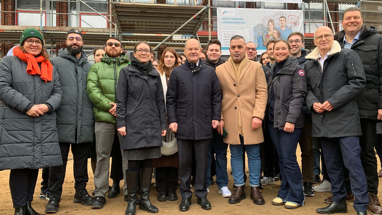 Das Bild zeigt eine Gruppe von Menschen meist mittleren Alters mit winterlichen Jacken, die draußen vor einen eingerüsteten Gebäude stehen.