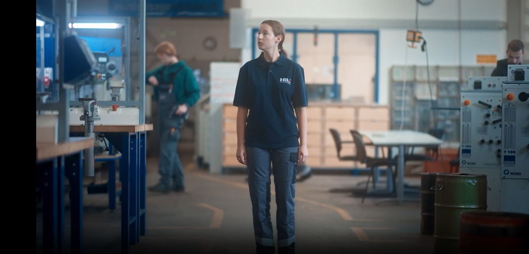 Das Foto zeigt eine junge blonde Frau, die in blauer Arbeitskleidung in einer Werkhalle steht.