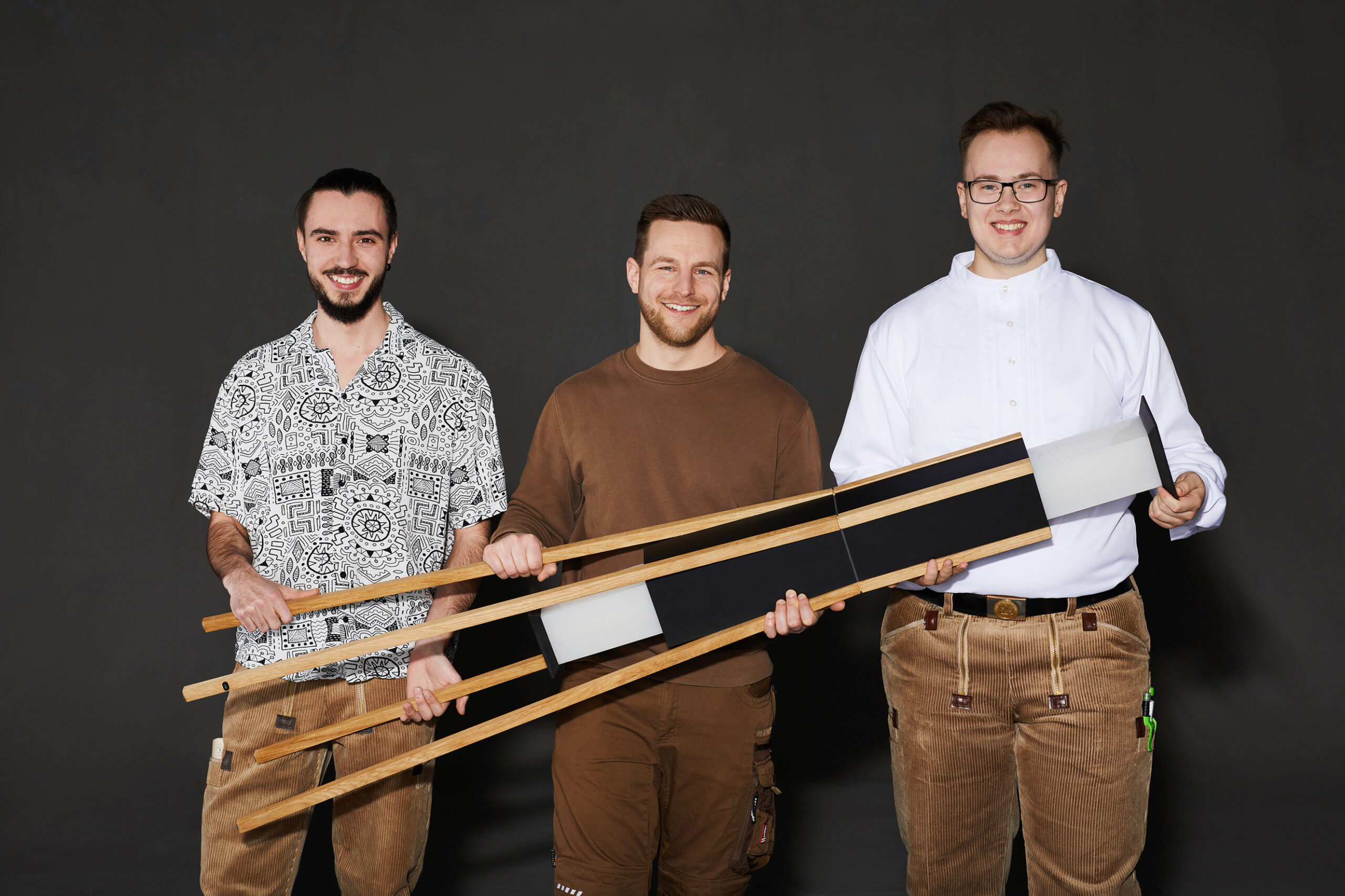 Drei junge Männer vor einem schwarzen Hintergrund. Sie halten eine Stehleuchte aus Holz in die Kamera und lächeln.