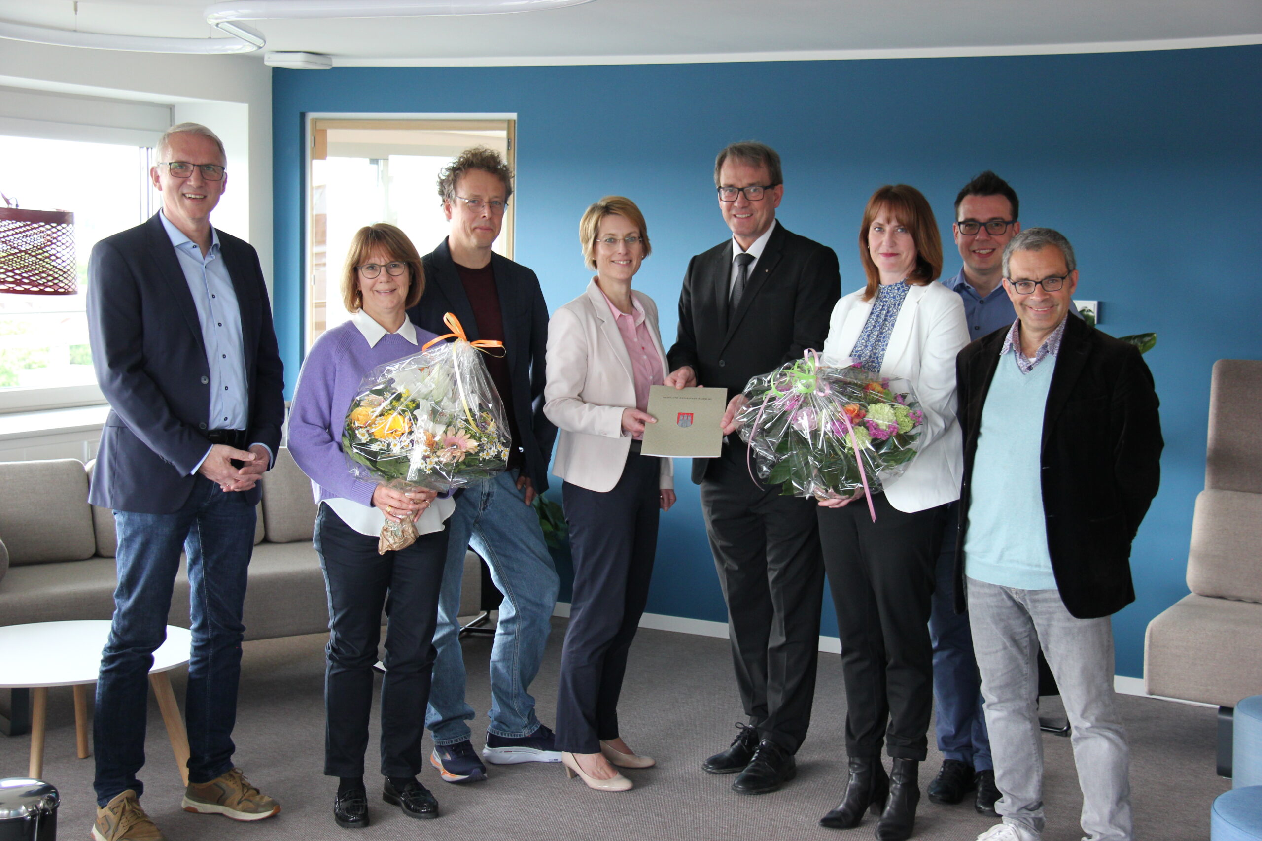 Drei Frauen und fünf Männer stehen vor einer blauen Wand und einem Sofa mit zwei Blumensträußen und einem Vertrag in der Hand. Sie schauen und lächeln in die Kamera.