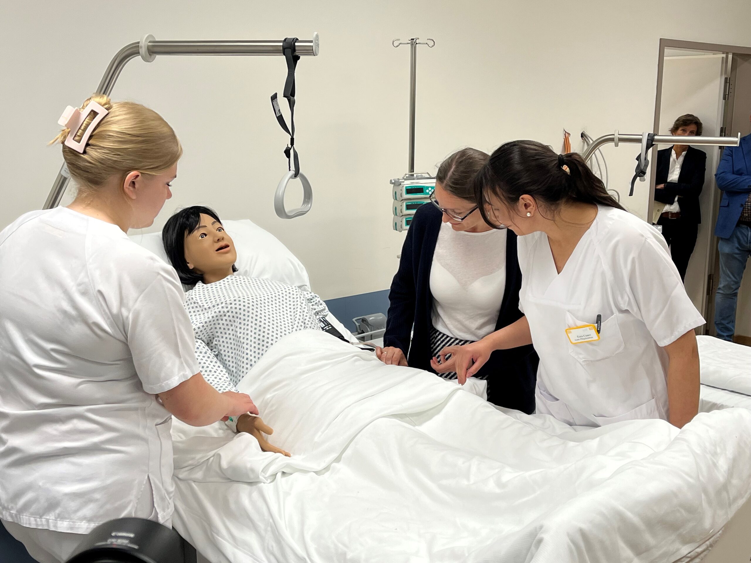 Das Foto zeigt 3 Frauen, davon 2 in weißen Krankenhauskittel und in der Mitte die Schulsenatorin Frau Bekeris, die sich über ein Krankenbett beuten, in dem eine menschengroße Puppe liegt.