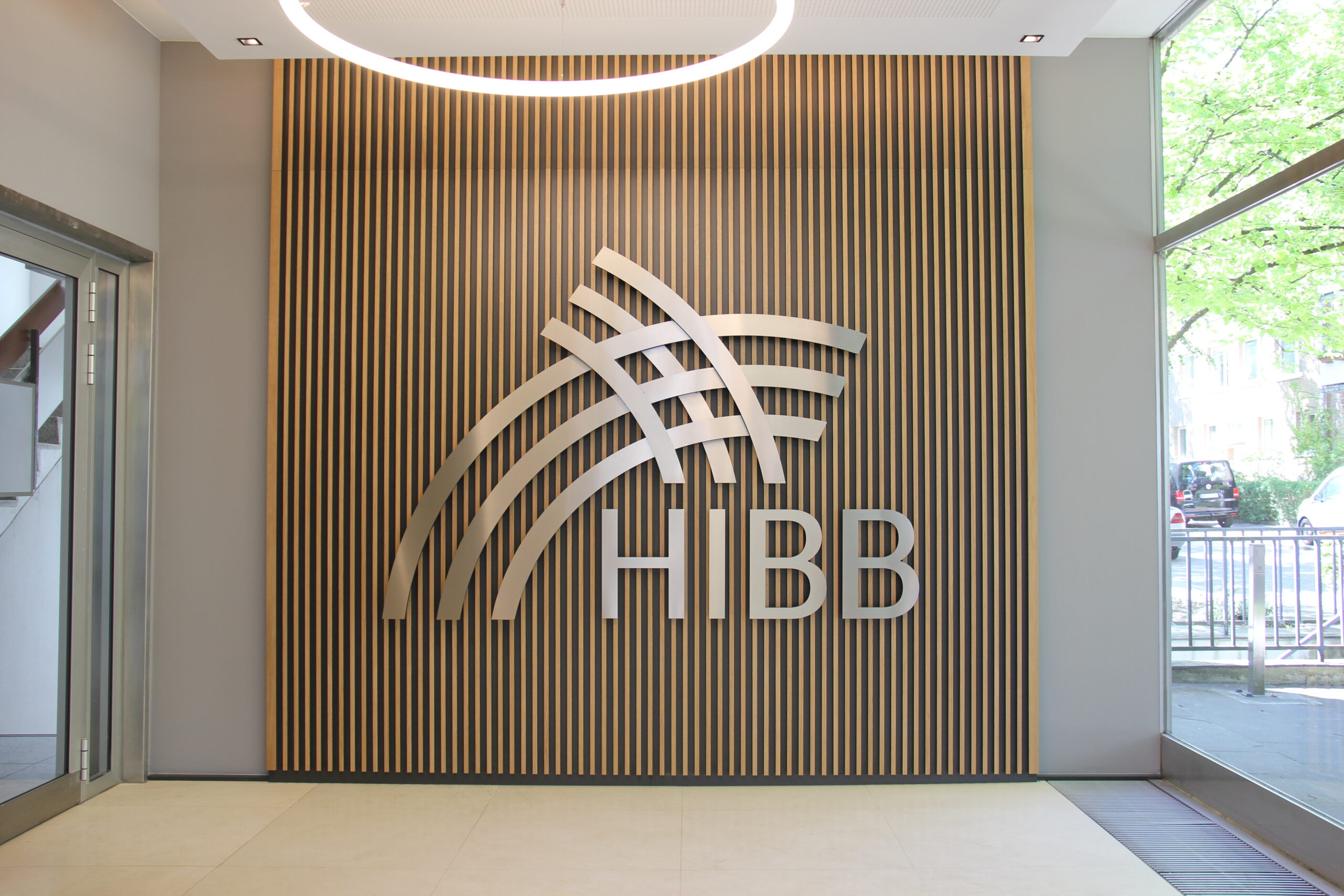 Das Foto zeigt den Eingangsraum des HIBB-Gebäudes. Man sieht eine Holzwand, auf der aus Metall das HIBB-Logo angebracht ist: Die Buchstaben H I B B und darüber 2 mal 3 sich überschneidende runde Linien.