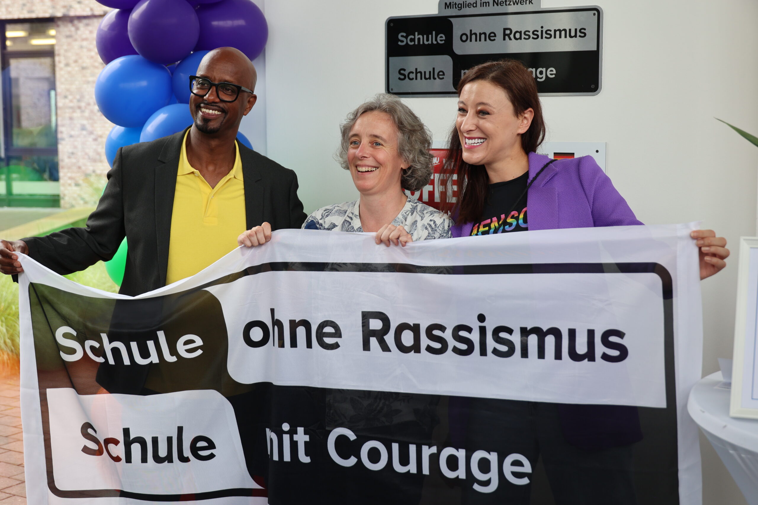 Ein Mann und zwei Frauen halten ein großes, schwarz-weißes Transparent hoch. Mit der Aufschrift "Schule ohne Rassismus - Schule mit Courage".