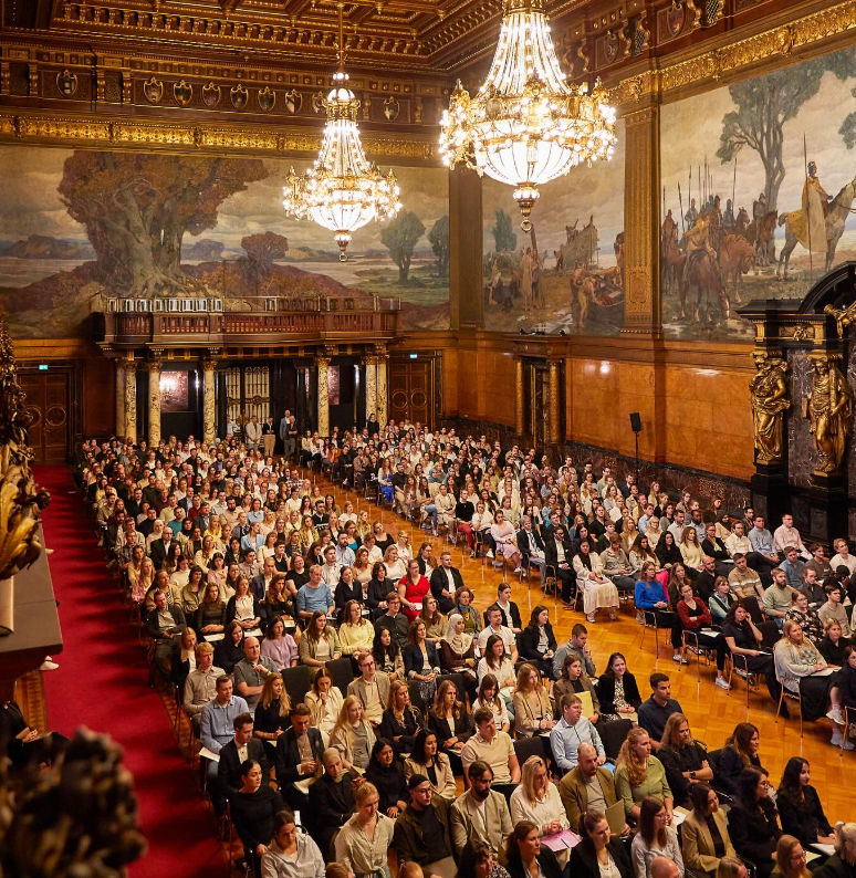 Ein großer Festsaal mit Kronleuchtern und Wandgemälden. Aus der Vogelperspektive. Unten sitzen zahlreiche junge Menschen.