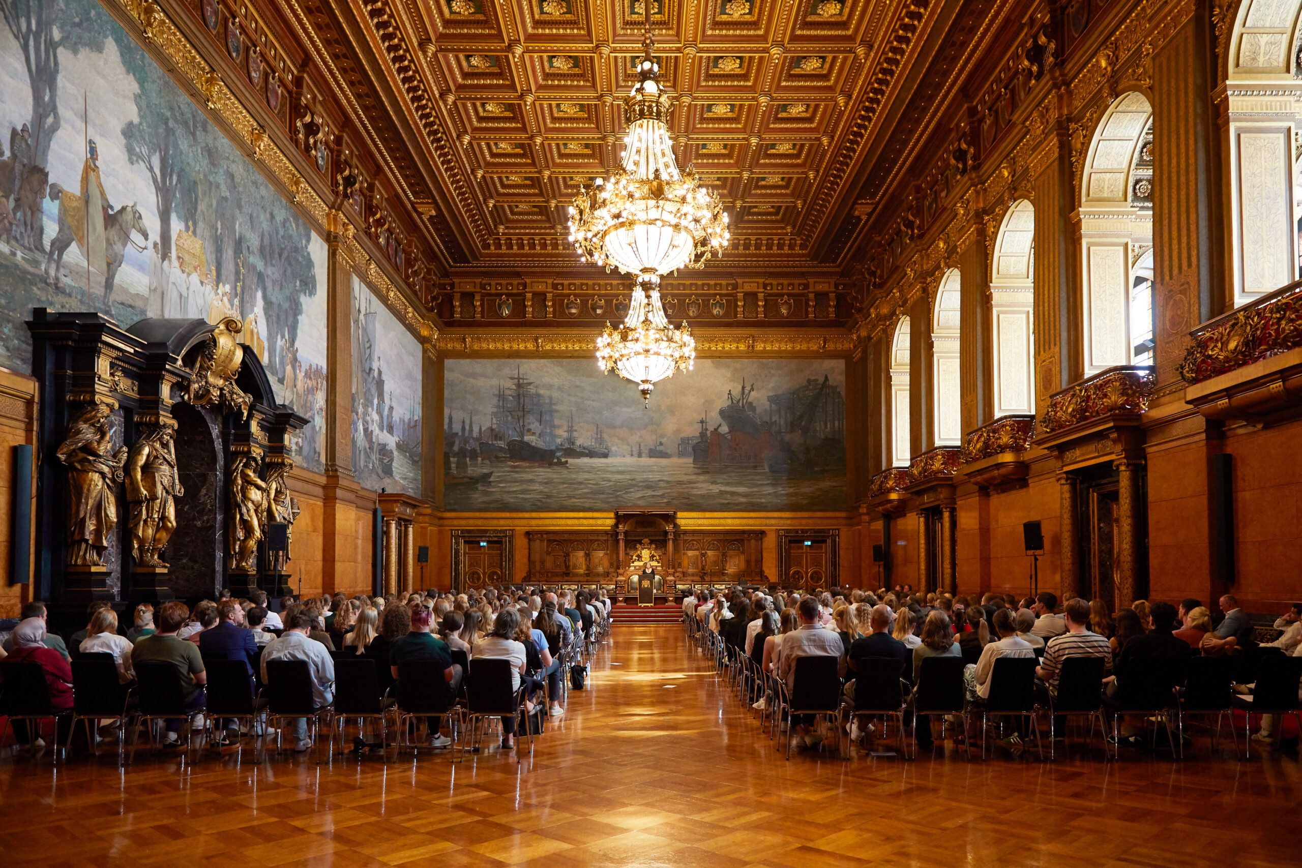Ein festlicher Saal mit Kronleuchtern und vielen Menschen auf Stühlen.
