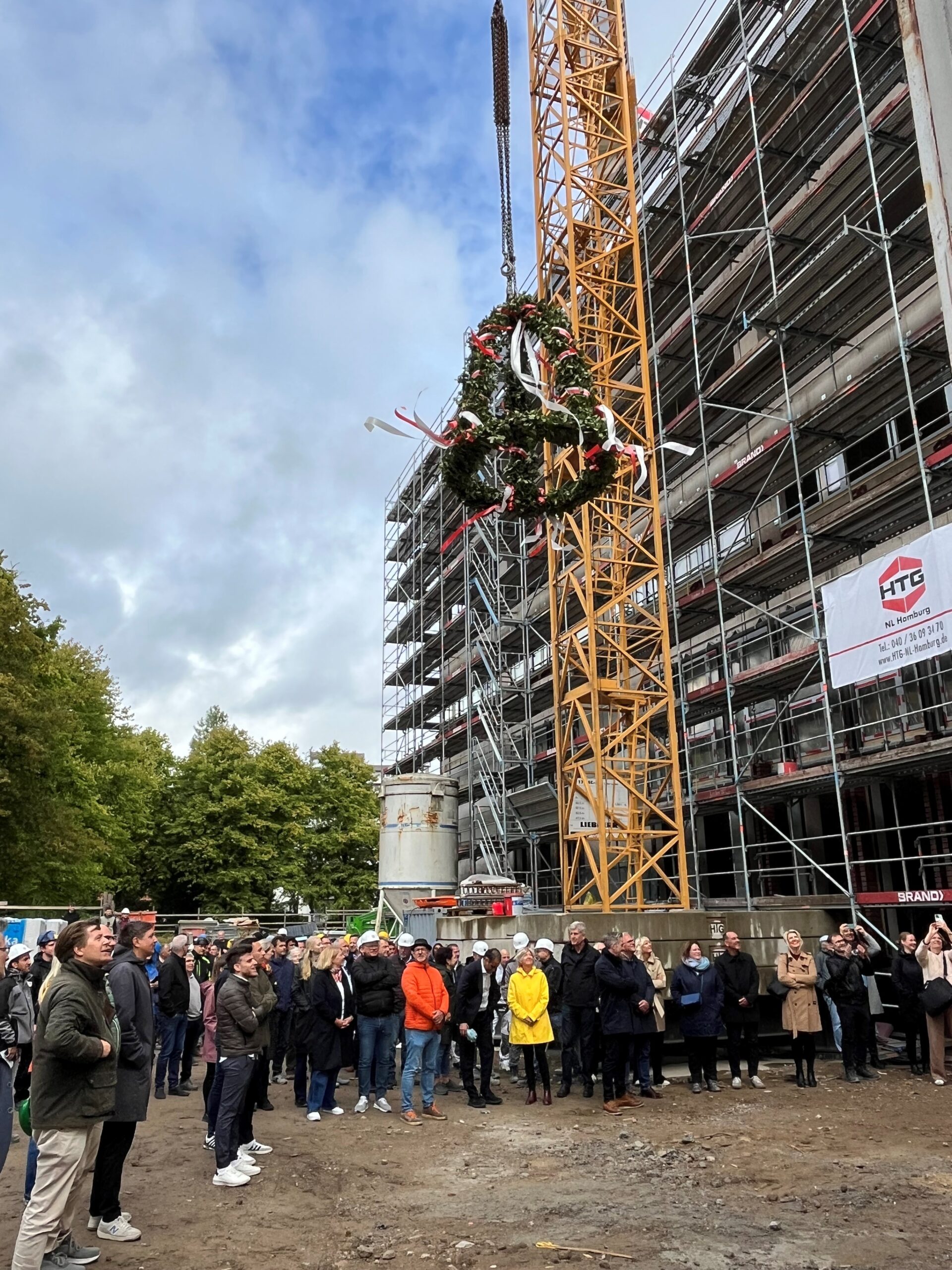 Vor einem großen Rohbau stehen viele Menschen und schauen nach oben zu einem Richtkranz.