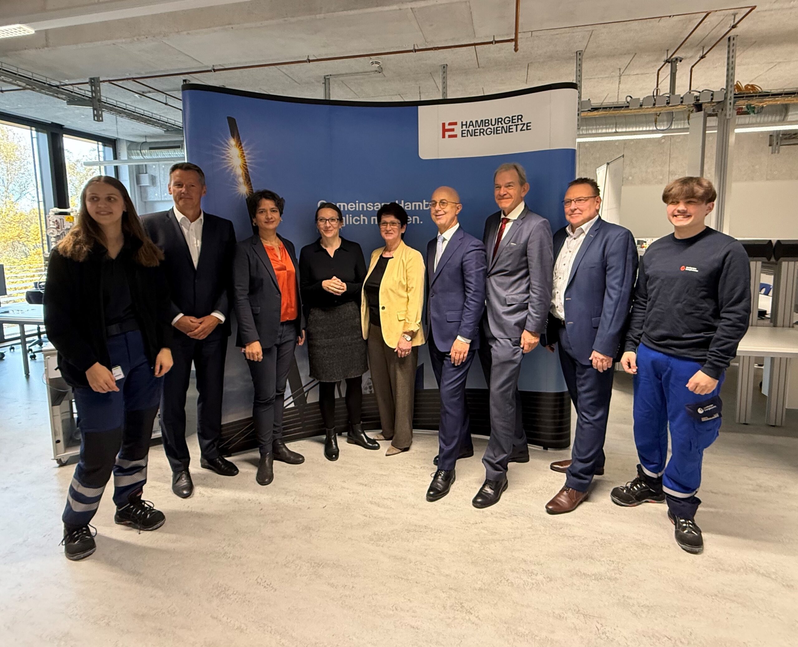 Neun Personen unterschiedlichen Alters stehen in einem Werkraum vor einer Leinwand, auf der "Hambrger Energienetze" steht,.